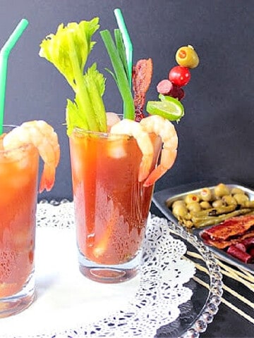 Two tall cocktail glasses filled with Bacon Bloody Mary along with shrimp, celery, scallions, and sausage.