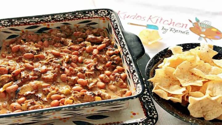 An offset horizontal photo of Cheesy Sausage Bean Dip in a square casserole dish and a bowl of nacho chips on the side.