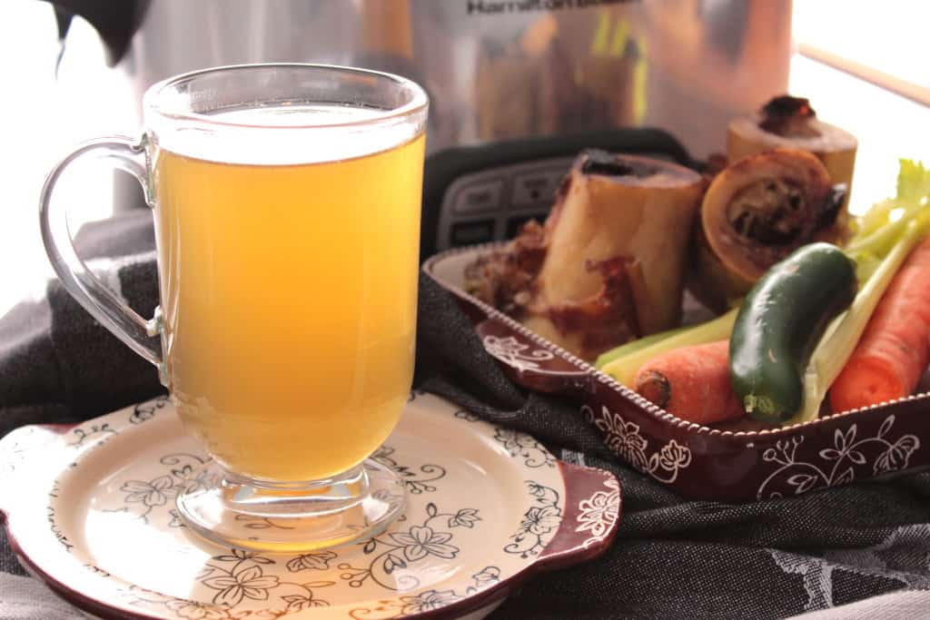 Homemade beef bone broth in a glass along with roasted bones and a slow cooker in the background for Soup, Stews, and Chowder Recipes.