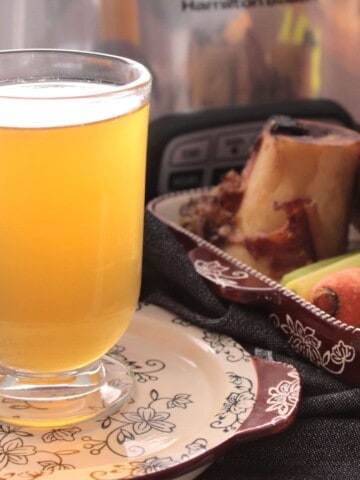 Homemade beef bone broth in a glass along with roasted bones and a slow cooker in the background.