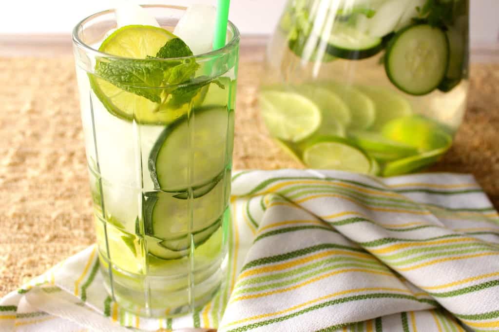 A horizontal photo of a glass of hydrating fitness water with sliced limes, cucumbers, and mint with ice and a green and yellow cloth napkin.
