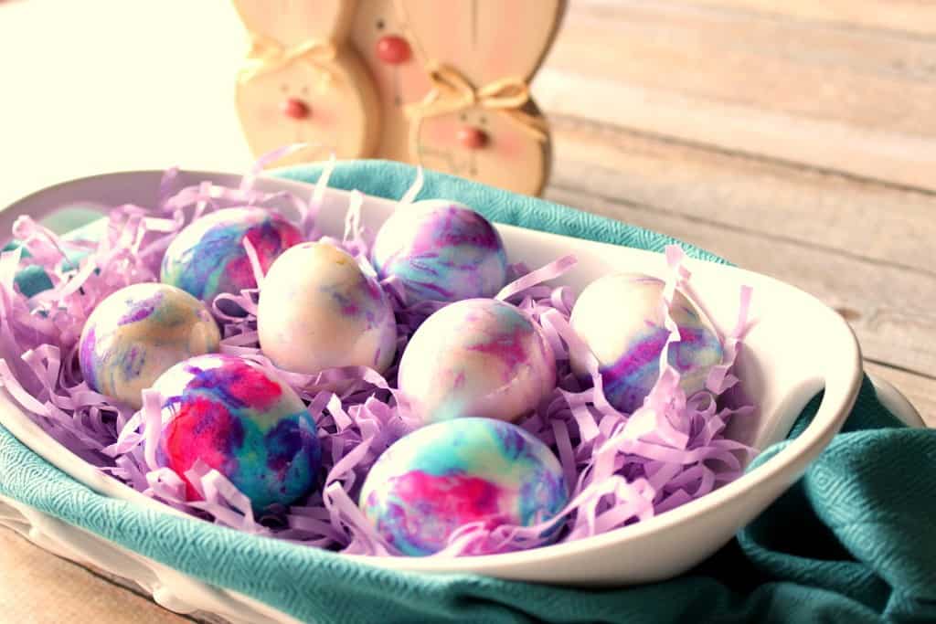 A horizontal photo of mayonnaise tie-dye eggs in a white bowl with purple paper and a blue napkin.