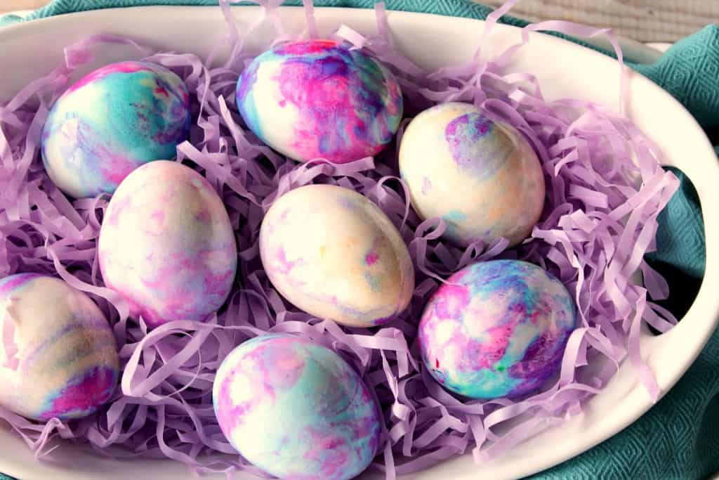Pretty Pastel Tie Dye Easter Eggs using Mayonnaise Overhead photo of pastel Easter eggs in a white bowl with purple paper shreds and a blue napkin.