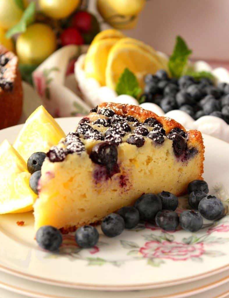 Slice of Fresh Lemon Ricotta Cake with Blueberries on a plate with lemon wedges and fresh blueberries.