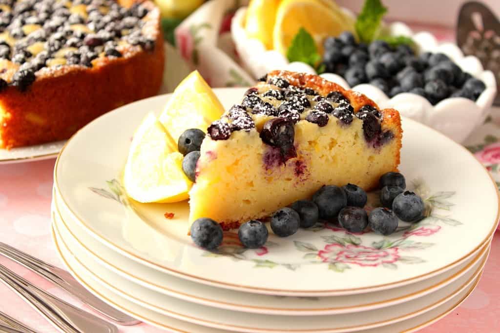 A slice of lemon ricotta cake on a stack of plates with a dish of blueberries and lemon slices in the background.