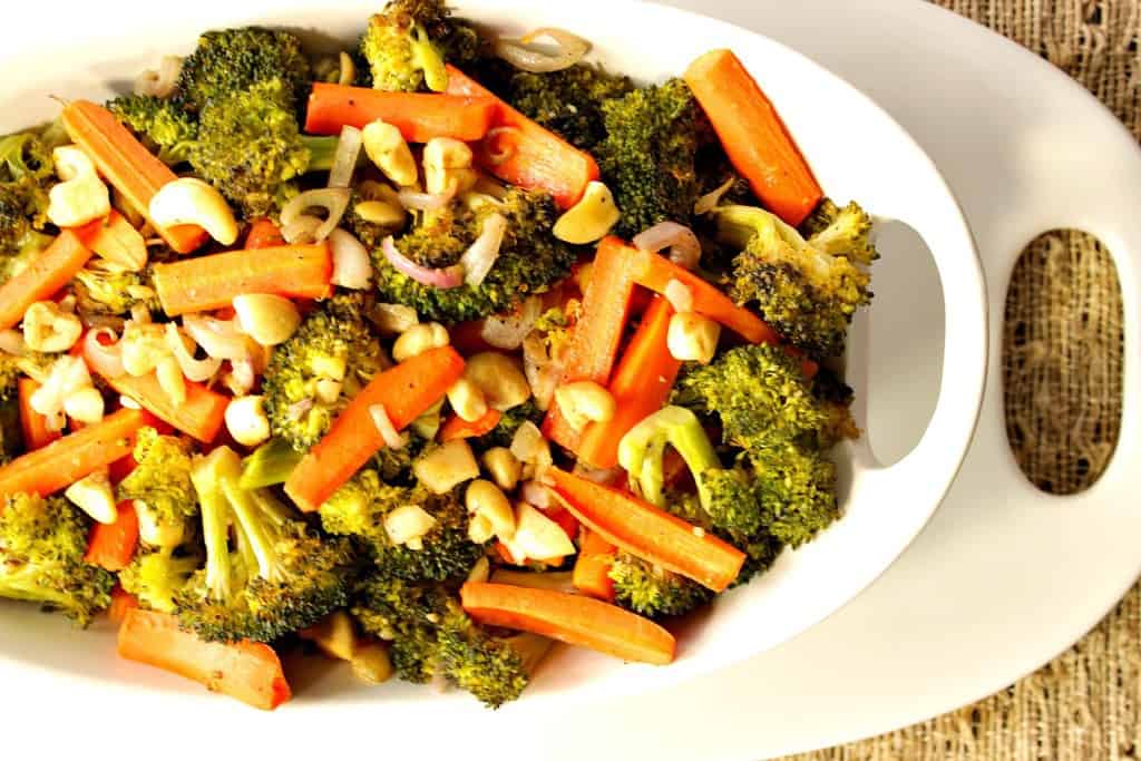 An overhead photo of Oven Roasted Carrots, Broccoli, Shallots and Cashews in a white oval bowl with a white oval platter underneath