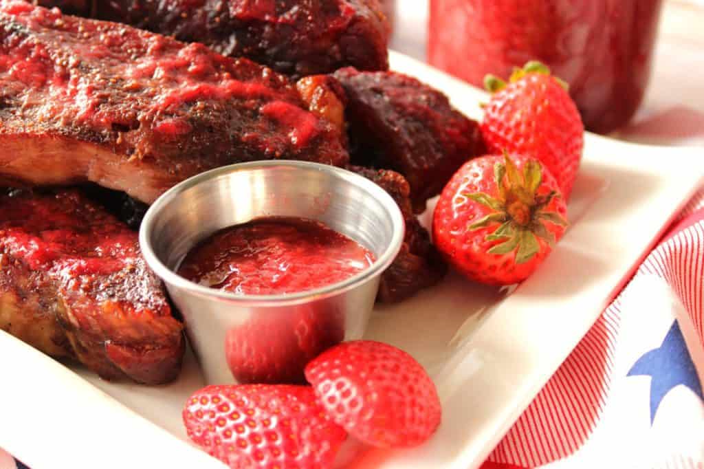 A closeup horizontal photo of a small dish of strawberry bbq sauce with ribs on a white plate and fresh strawberries.