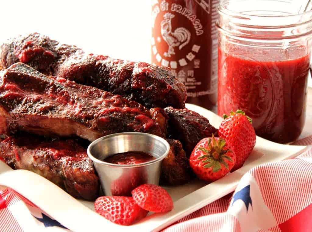 A horizontal photo of a pile of pork ribs on a white plate the strawberries and bbq sauce.