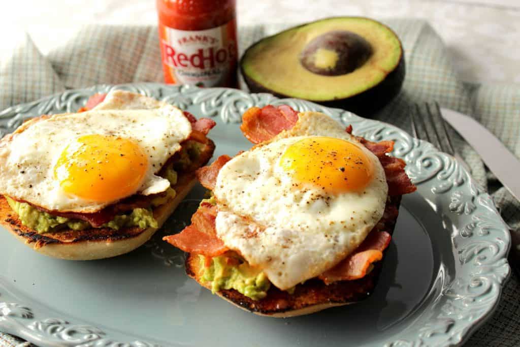 A horizontal photo of two avocado toast breakfast sandwiches on a blue plate with sunny side up eggs and bacon.