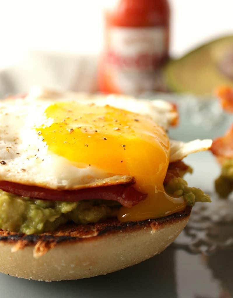 A closeup vertical photo of a runny yolk egg on an avocado toast breakfast sandwich with bacon.