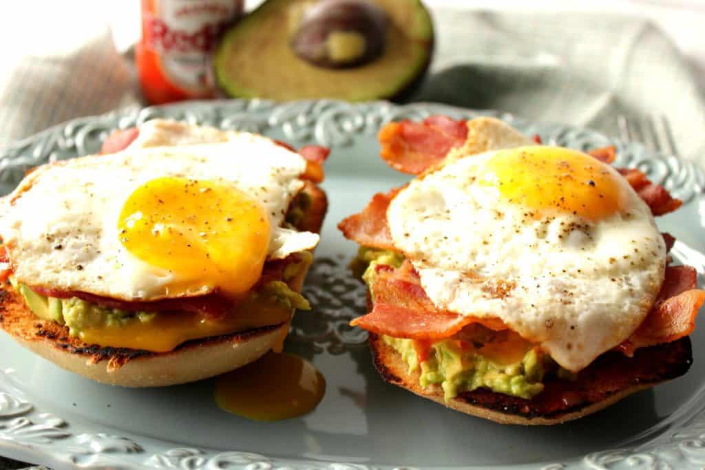 Two avocado toast with bacon and egg breakfast sandwiches on a blue plate with an avocado half and hot sauce in the background.