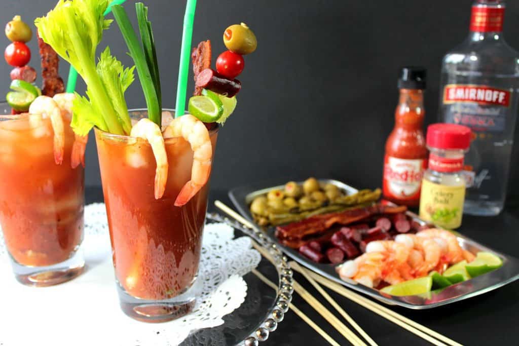 2 bloody Mary drinks on a tray with a doily, loaded with shrimp, celery, olives, and straws.