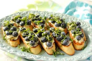 A blue platter filled with bruschetta topped with fresh blueberries and fresh basil.