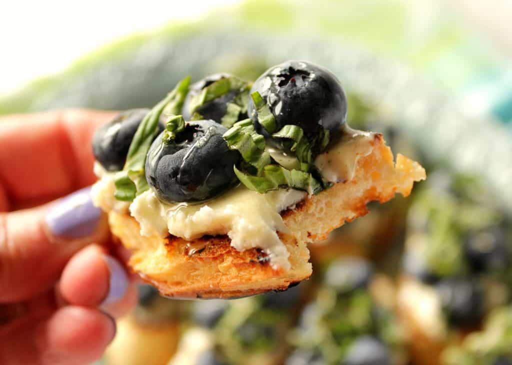 A closeup photo of a hand holding a blueberry basil bruschetta with a bite taken out.