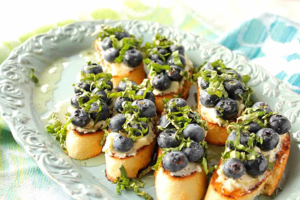 Blueberry basil bruschetta on a blue plate with fresh blueberries, goat cheese, and fresh basil