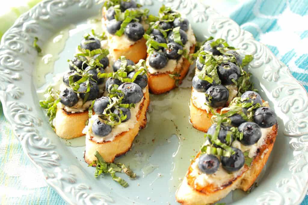 A blue platter with some blueberry basil bruschetta missing, along with a drizzle of honey, and some fresh basil over top