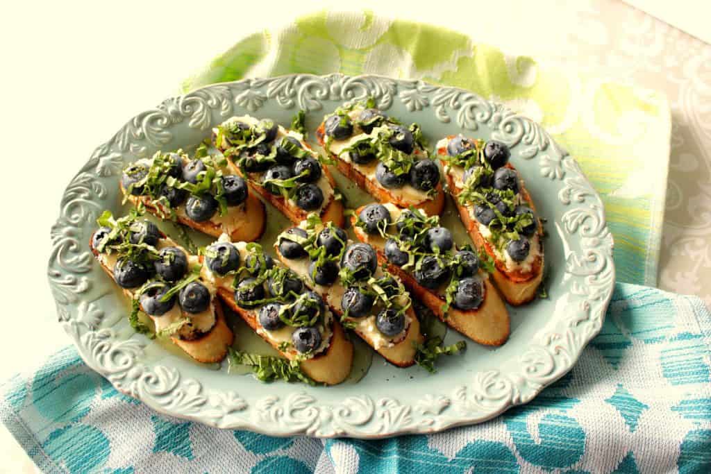On overhead photo of a plate of goat cheese and blueberry basil brushchetta with a blue napkin