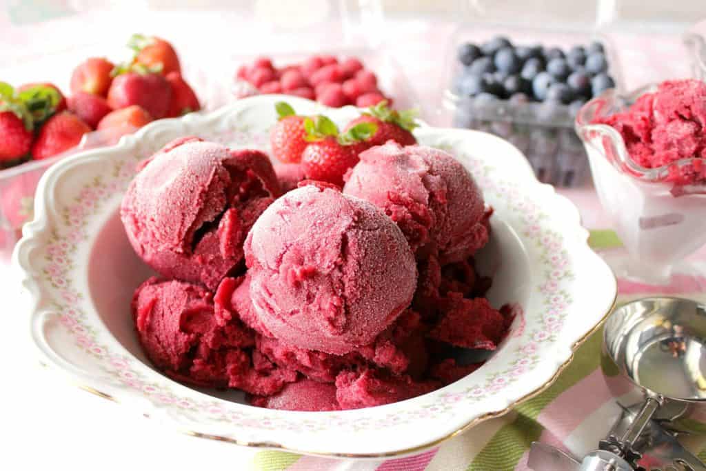 Pretty bowlful of pink sorbet with berries in the background and an ice cream scoop.