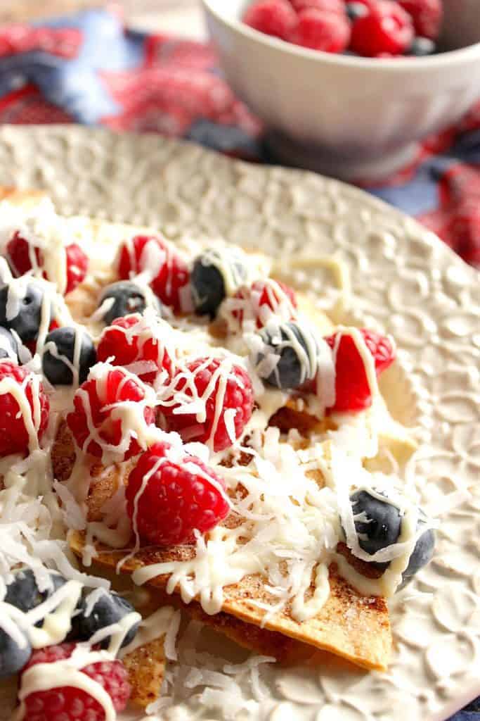 Vertical closeup image of crispy cinnamon sugar dessert nachos with red and blue berries and a white chocolate and coconut drizzle.