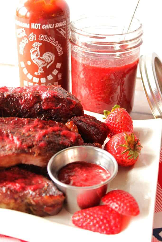 A closeup vertical photo of ribs on a plate with strawberry bbq sauce in the background.