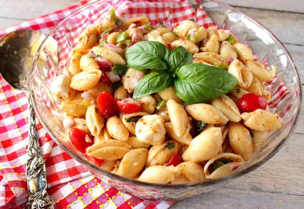 Red and white checkered napkin with a large serving spoon and a large glass bowl of shell pasta salad.