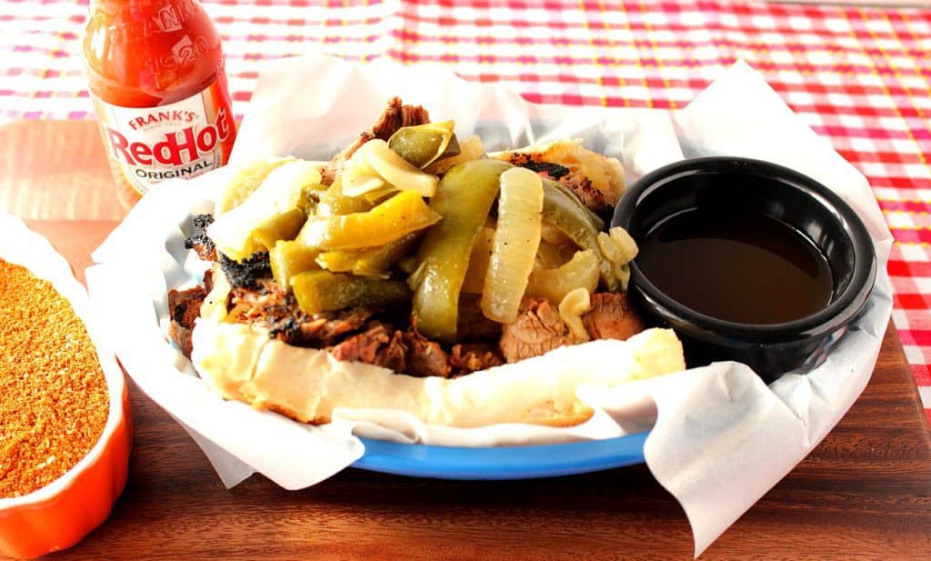 A skirt steak sandwich in a blue takeout basket with a small container of beef juice and a bottle of hot sauce in the background.