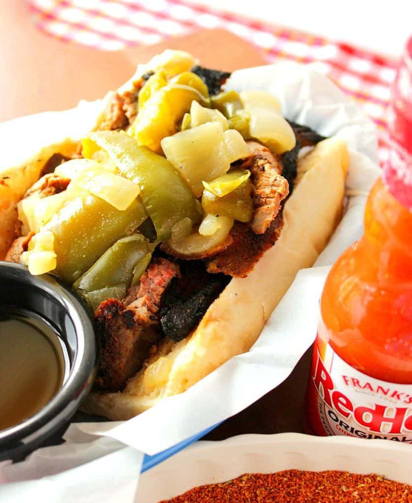 Closeup image of a dry rub skirt steak sandwich with sauteed green peppers and onions and a bottle of hot sauce on the side.