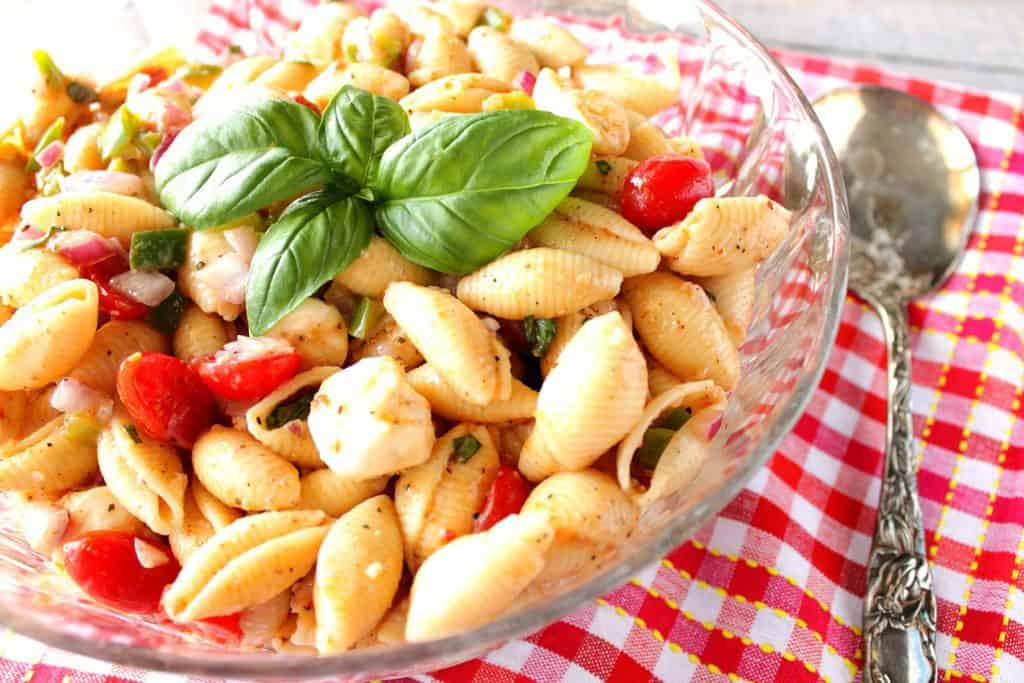 Large glass bowl filled with shell pasta salad, grape tomatoes and fresh basil leaves.