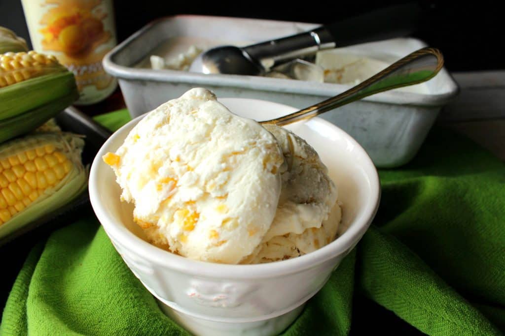 A small white bowl filled with no-churn roasted sweet corn ice cream with a spoon and a ice cream scoop in the background