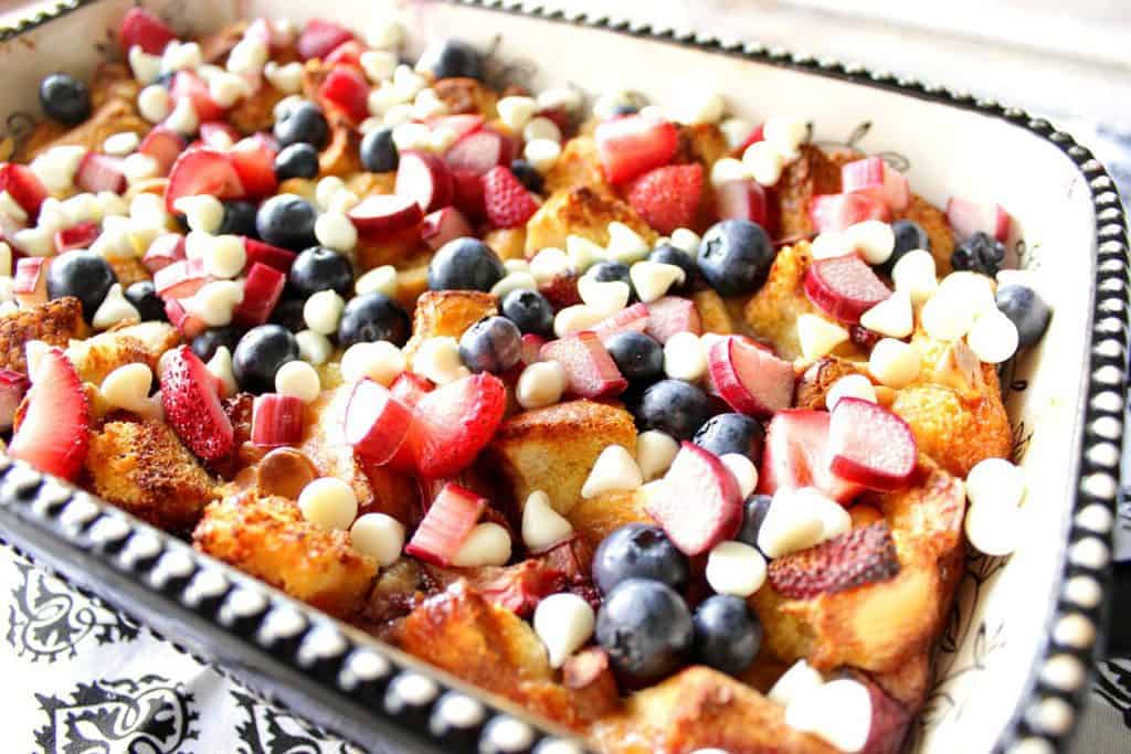 Closeup photo of bread pudding in a casserole dish with strawberries, rhubarb, blueberries and white chocolate chips.