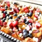 Closeup photo of bread pudding in a casserole dish with strawberries, rhubarb, blueberries and white chocolate chips.