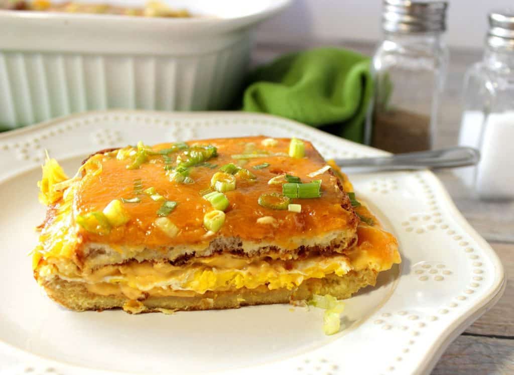 A slice of Egg Sandwich Casserole on a white plate with scallions and cheddar cheese.