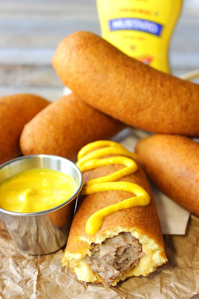 A closeup vertical image of a bratwurst on a stick with a bite taken out and with yellow mustard on the side and top.