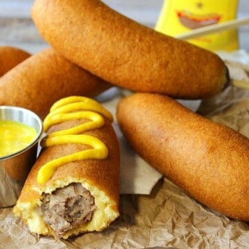 A pile of beer batter bratwurst on a brown paper bag with mustard and a bite taken out
