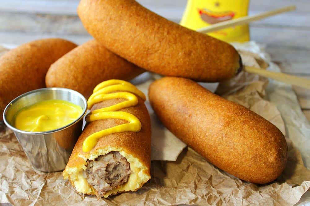 A pile of beer batter bratwurst on a brown paper bag with mustard and a bite taken out