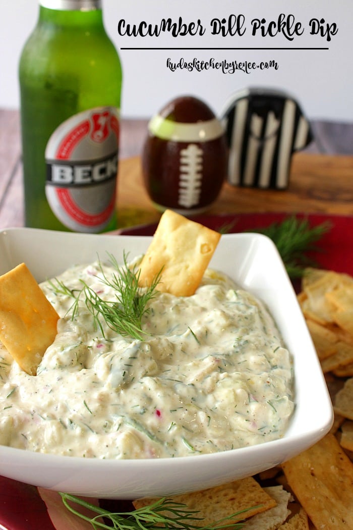 This Flavor Packed Cucumber Dill Pickle Dip will keep you coming back for more. It's big bold flavor is great for chips, crackers and even vegetables. - Kudos Kitchen by Renee - www.kudoskitchenbyrenee.com Closeup photo of cucumber dill dip with crackers and fresh dill in a square white bowl.