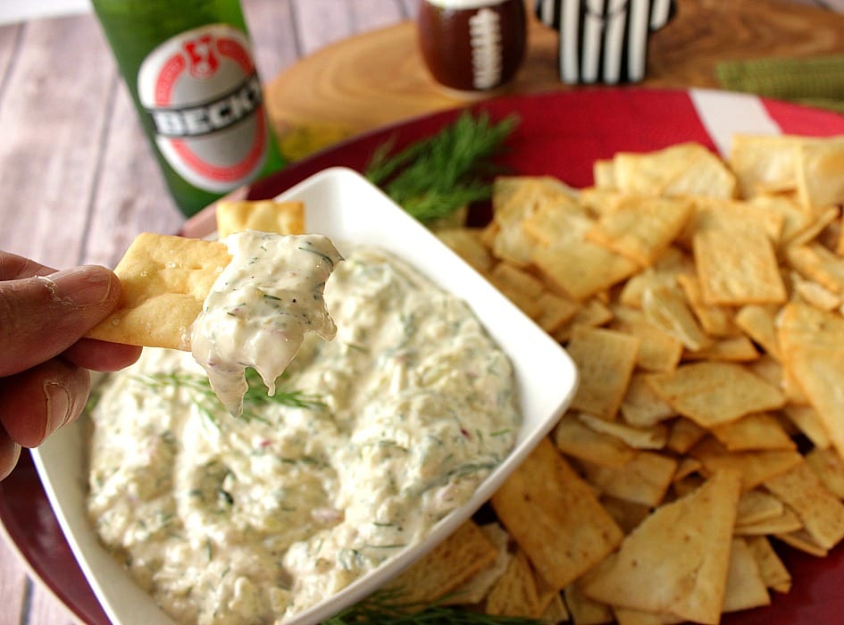 A cracker with Cucumber Dill Pickle Dip along with a bottle of beer in the background and crackers on a platter.