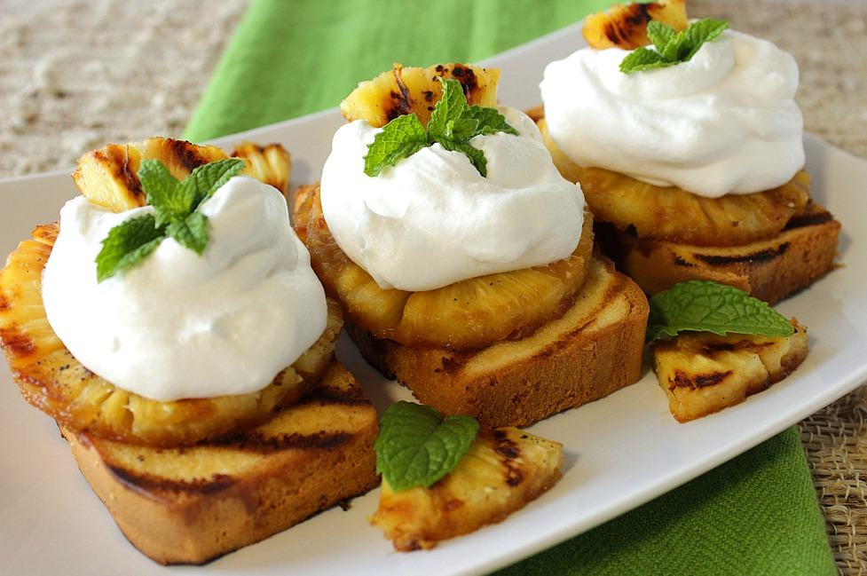 Three slices of grilled pound cake topped with grilled pineapple and whipped cream on a white plate with a green napkin and mint.