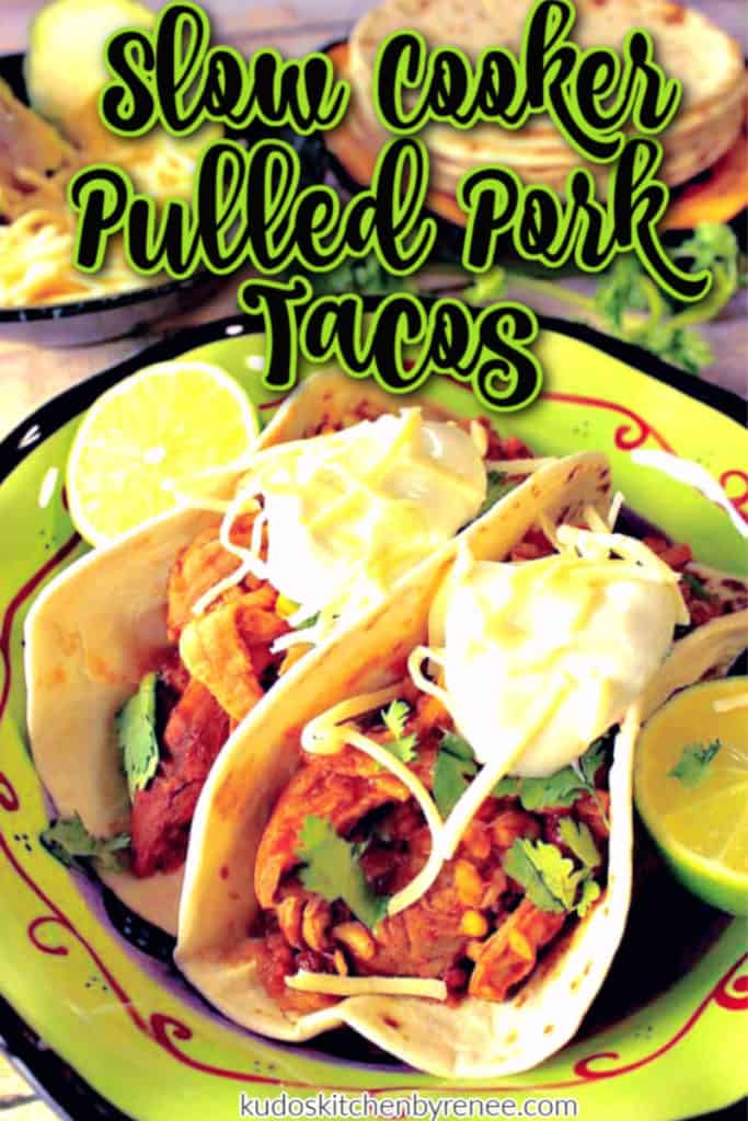 Closeup vertical image of two slow cooker pulled pork tacos in a green bowl with sour cream, cilantro, and shredded cheese.