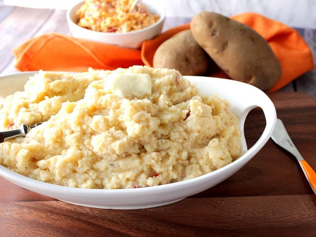 A white oval bowl filled with pimento cheese mashed potatoes with butter on top and Idaho potatoes and an orange napkin in the background.