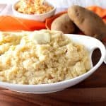 A white oval bowl filled with pimento cheese mashed potatoes with butter on top and Idaho potatoes and an orange napkin in the background.