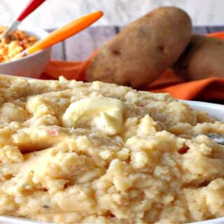 A white oval bowl filled with Pimento Cheese Mashed Potatoes along with some russet potatoes in the background.