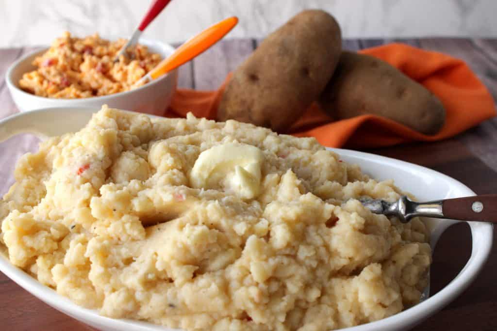 A horizontal photo of a white oval bowl filled with Pimento Cheese Mashed Potatoes along with a spoon and russet potatoes in the background