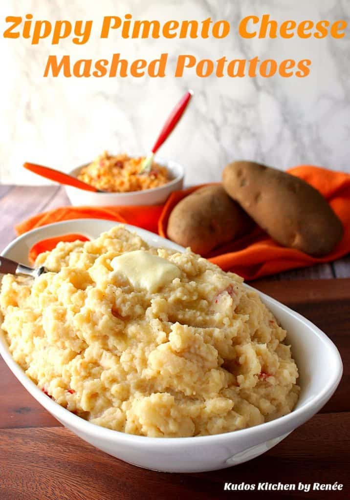 A vertical photo of a bowl of Pimento Cheese Mashed Potatoes along with a small bowl of pimento cheese in the background along with russet potatoes.