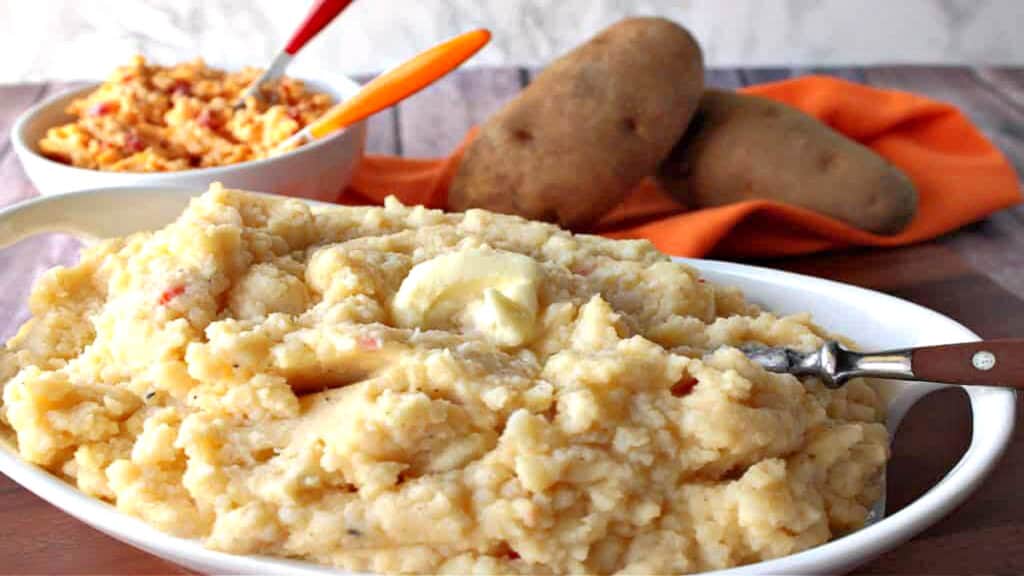 A white oval bowl filled with Pimento Cheese Mashed Potatoes along with some russet potatoes in the background.