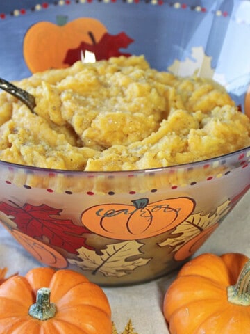 A glass bowl filled with Pumpkin Mashed Potatoes along with baby pumpkins in the foreground.
