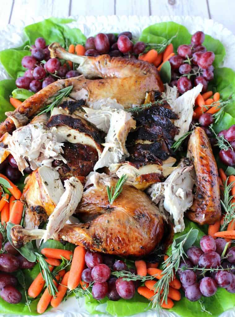 A vertical photo of a colorful platter of a balsamic herb roast turkey which is sliced and surrounded by grapes, carrots, herbs, and greens.