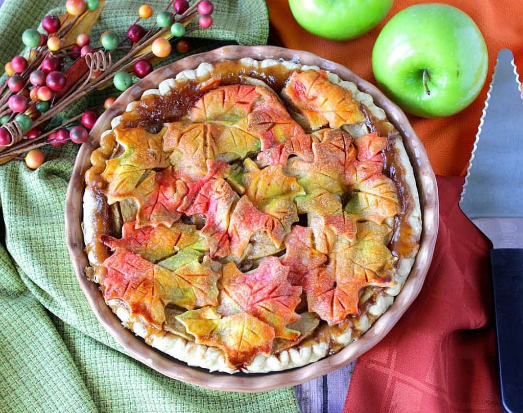Overhead photo of a pie pan with an autumn leaves apple pie, colorful napkins, a pie server, and apples.