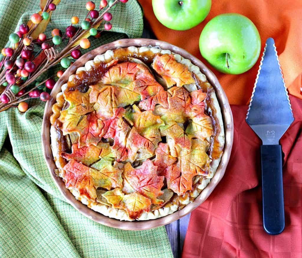 Overhead photo of an autumn leaves apple pie with a pie server and apples.
