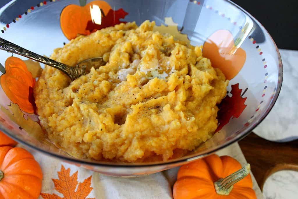 An offset horizontal photo of a bowl full of orange Pumpkin Mashed Potatoes with a serving spoon and melted butter.